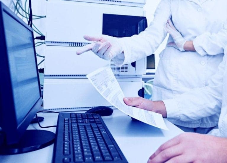 High-tech pharmaceutical laboratory with scientists discussing data on a large monitor, safety equipment, and chemical reagents.